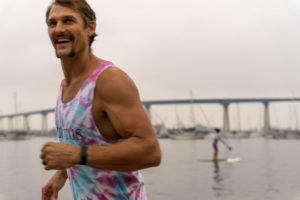 Man with mustache in tie-die muscle shirt running on beach with a huge smile.