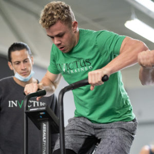 An Invictus coach looks over the shoulder of an athlete who is standing on the Assault Bike while pedaling.