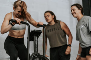 Two female athletes cheer on a third who is riding the Assault Bike so hard that her hair is blowing everywhere.