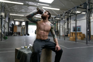 Male athlete sitting on a box while drinking from a shaker bottle.