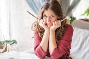 Woman with fork and knife in hands is sitting with her elbows on the table and a questioning look on her face.