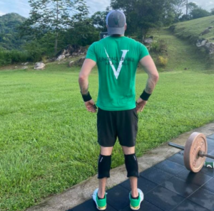 Male athlete on a driveway standing next to a barbell whilst gazing across his lawn.